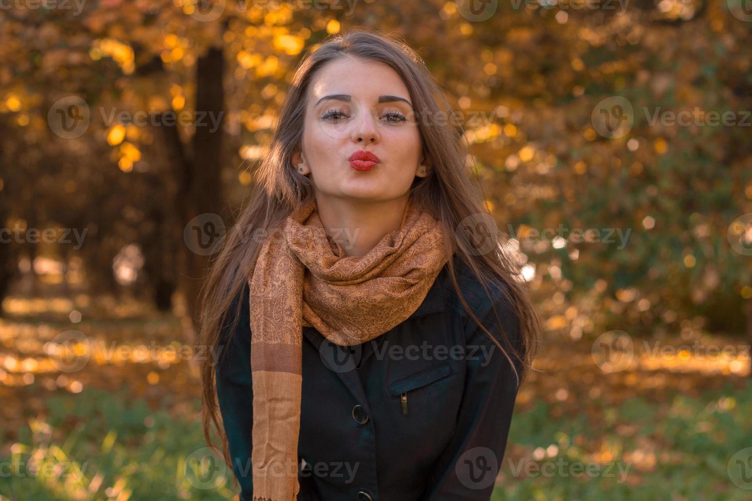 schönes Mädchen steht im Herbstpark in einem langen Schal, der direkt schaut und Kuss sendet foto