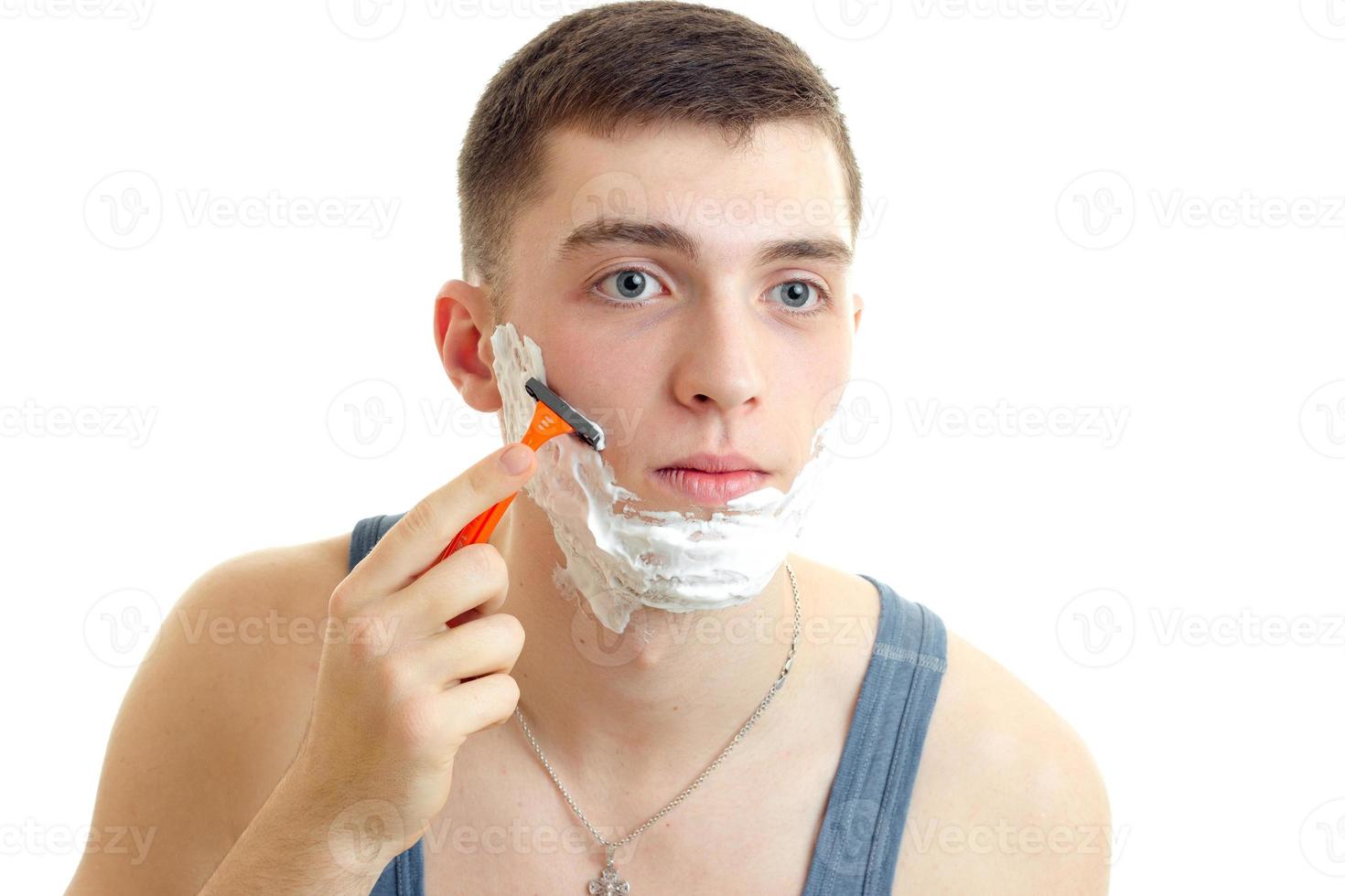 Der junge Mann schaut nach vorne und rasiert seinen Bart mit einem Rasiermesser mit Schaum foto