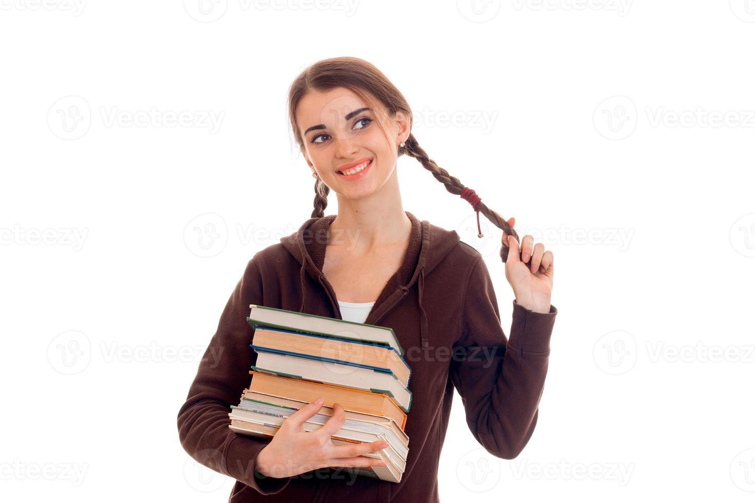 Fröhliches, intelligentes Studentenmädchen in brauner Sportkleidung mit vielen Büchern in der einen Hand, die ihr Haar berührt und isoliert auf weißem Hintergrund lächelt foto