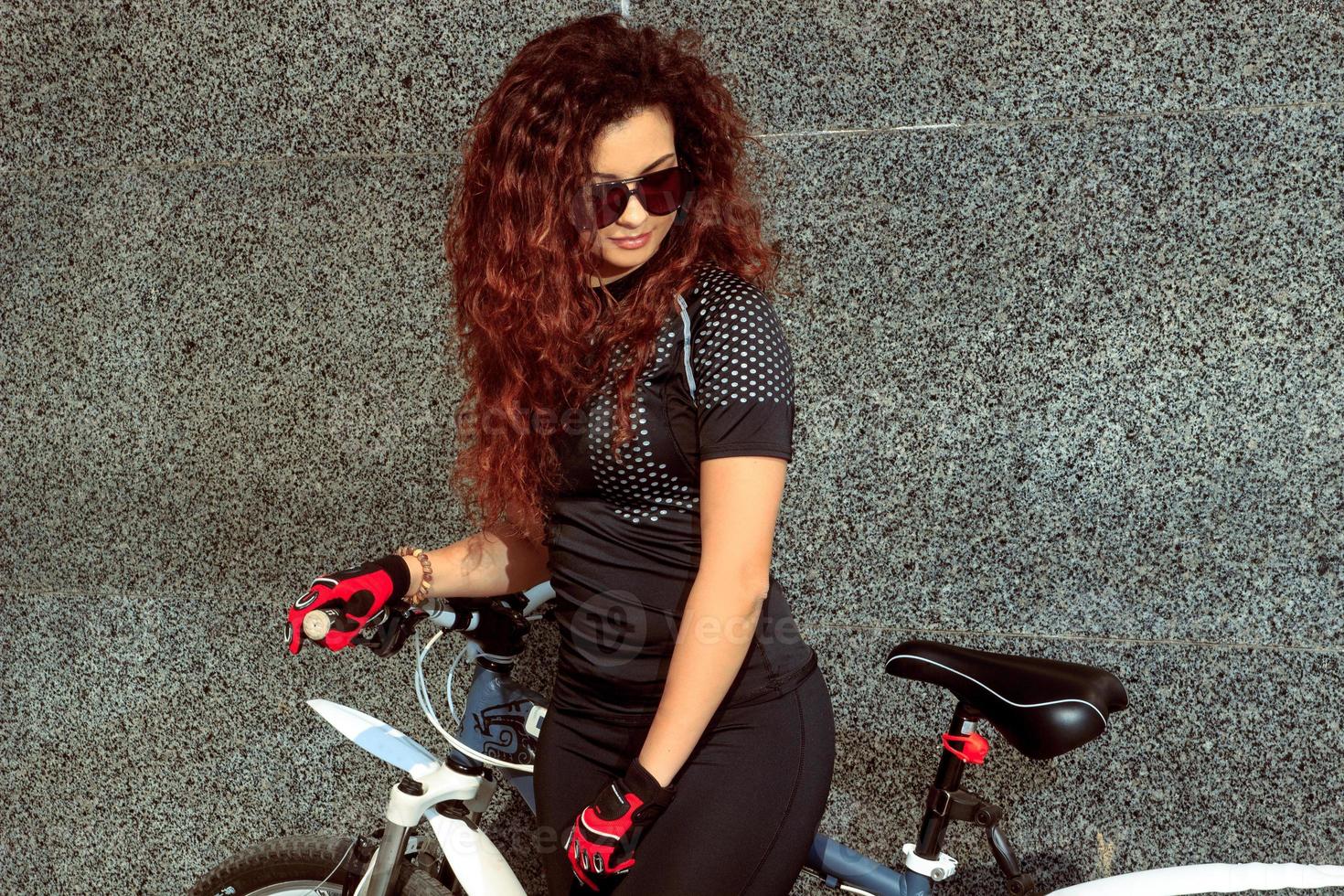 Glamour-Frau mit Sonnenbrille mit lockigem rotem Haar auf dem Fahrrad foto
