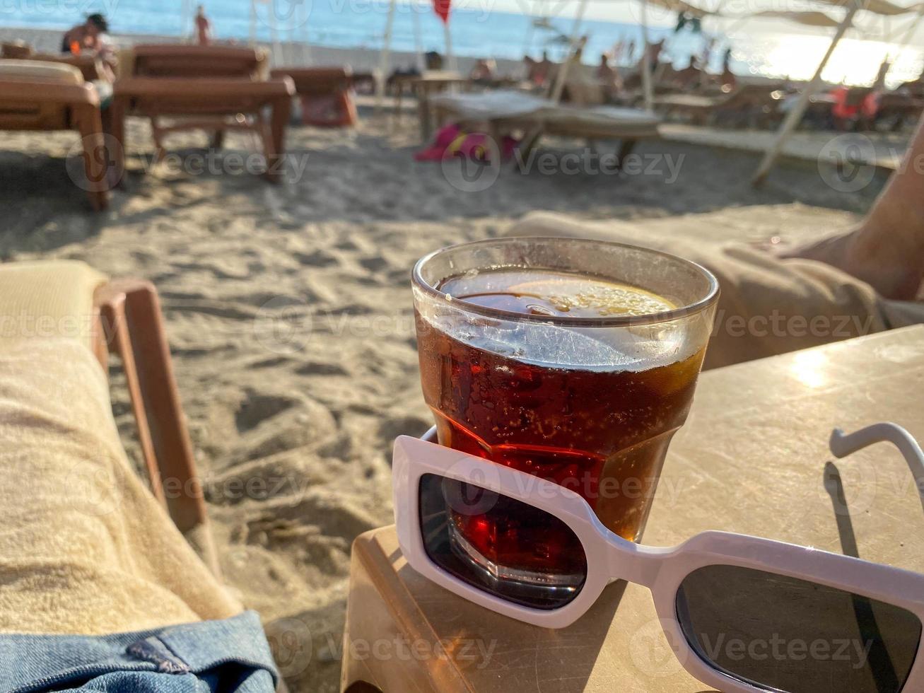 ein glas mit einem alkoholfreien getränk brauner cocktail mit soda und whisky und sonnenbrille auf einem tisch am strand am meer in einem hotel im urlaub in einem paradiesischen warmen osttropischen landresort foto