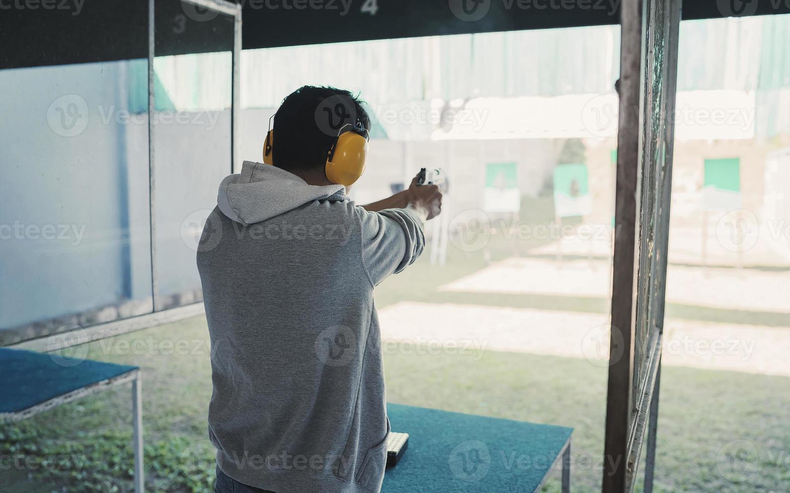 asiatischer schütze, der geräuschunterdrückende kopfhörer und schwarze kleidung trägt, die das schießen einer kurzen waffe auf dem schießstand üben. Schießsport zur Meditation und Selbstverteidigung, Freizeitaktivitäten. foto