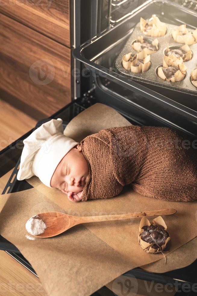 neugeborenes baby mit kochmütze liegt mit muffins auf dem ofenblech foto