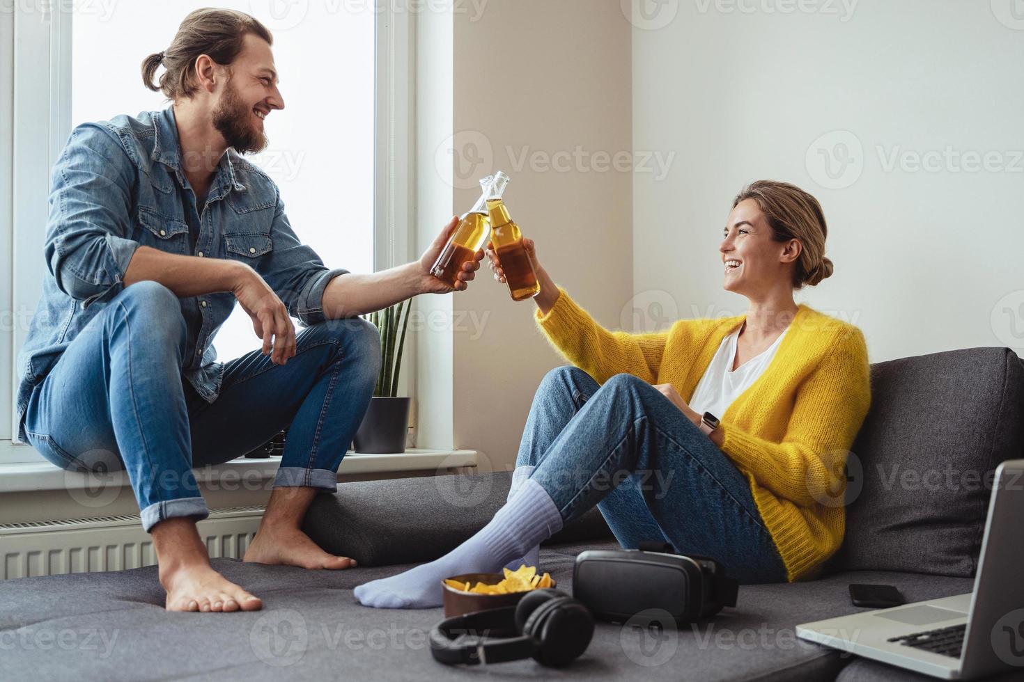 junges fröhliches paar, das auf dem sofa sitzt, bier trinkt und nachos isst foto