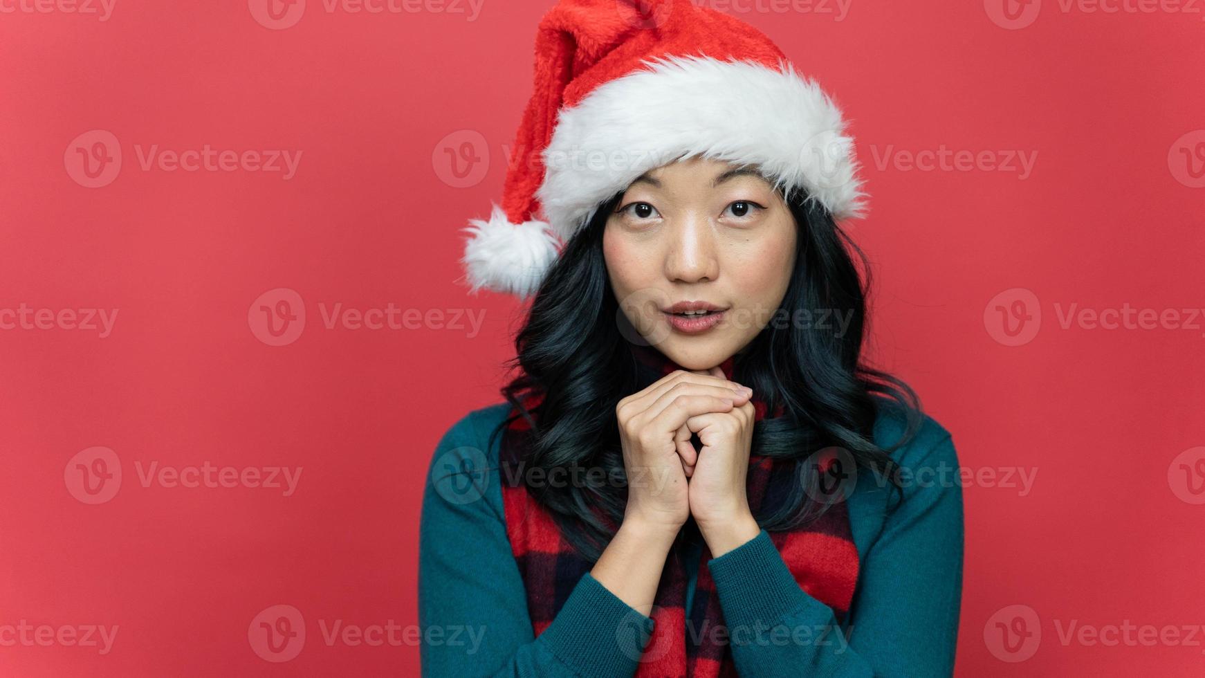 Nahaufnahme des Porträts einer jungen asiatischen Frau mit langen Haaren, die eine Weihnachtsmütze trägt und sich einen Wunsch isoliert über rotem Hintergrund macht. Emotion Lächeln foto