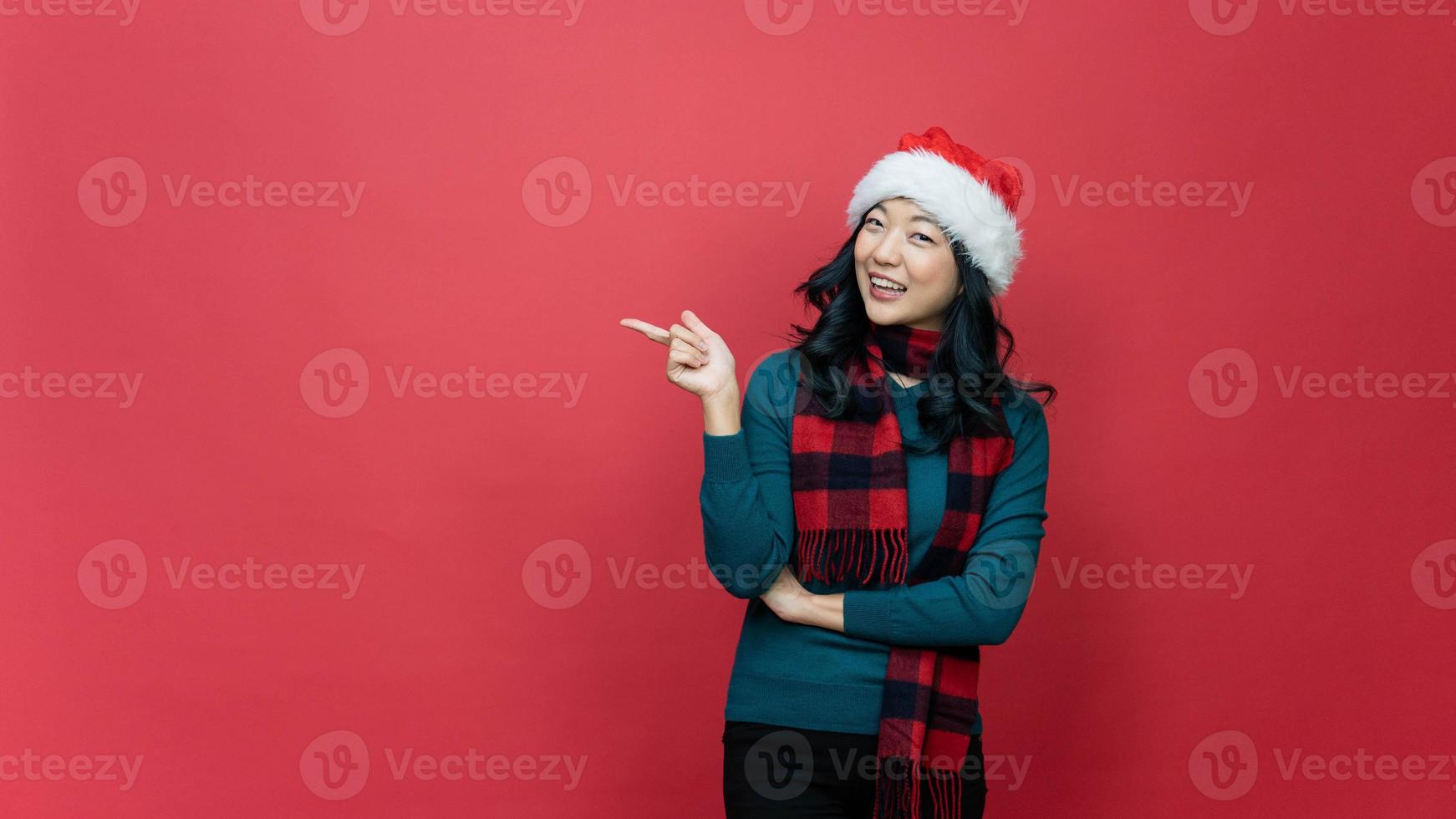 Weihnachtsverkaufskonzept. glückliche junge frau, die auf leeren raum auf rotem studiohintergrund, panorama zeigt. hübsche dame in weihnachtsmütze und warmem pullover macht weihnachtsaktion oder sonderangebot foto