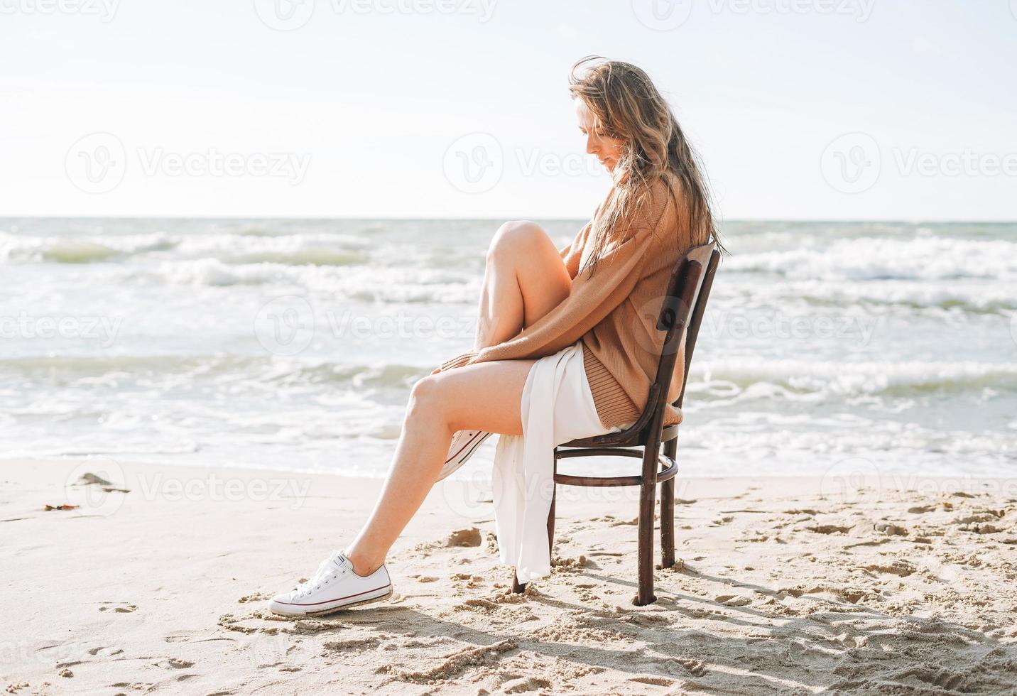 junge sorglose schöne Frau mit langen Haaren im Pullover, die auf einem Stuhl am Meeresstrand sitzt foto