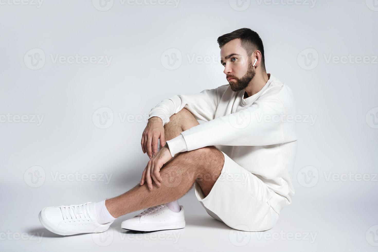 Gutaussehender Mann mit weißem Sweatshirt und Shorts mit drahtlosen Ohrstöpseln im Sitzen foto