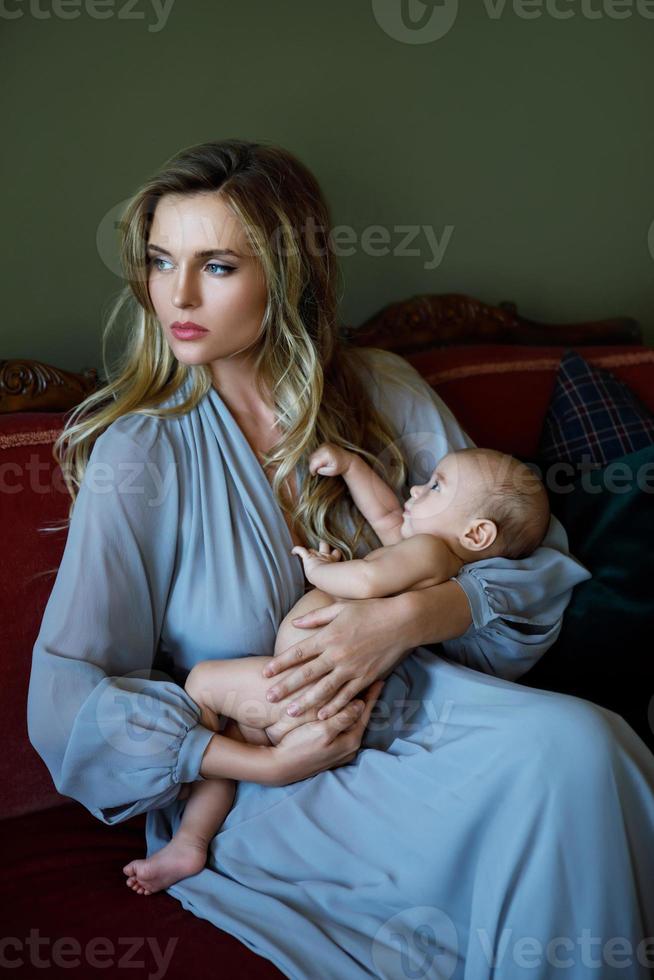 junge schöne mutter, die ein graues luxuskleid trägt, das ihr baby an den händen hält foto