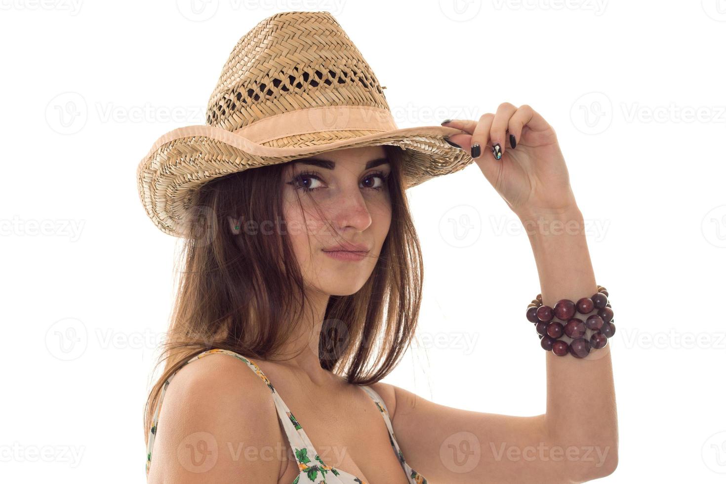 süßes junges brünettes Mädchen in Sommerkleidung mit Blumenmuster und Strohhut, das isoliert auf weißem Hintergrund in die Kamera schaut foto