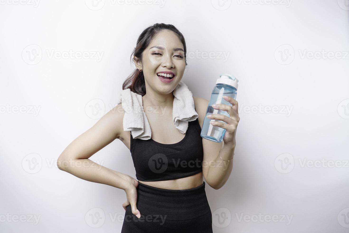 sportliche asiatin posiert mit einem handtuch auf der schulter und hält eine flasche wasser, lächelt und entspannt sich nach dem training foto