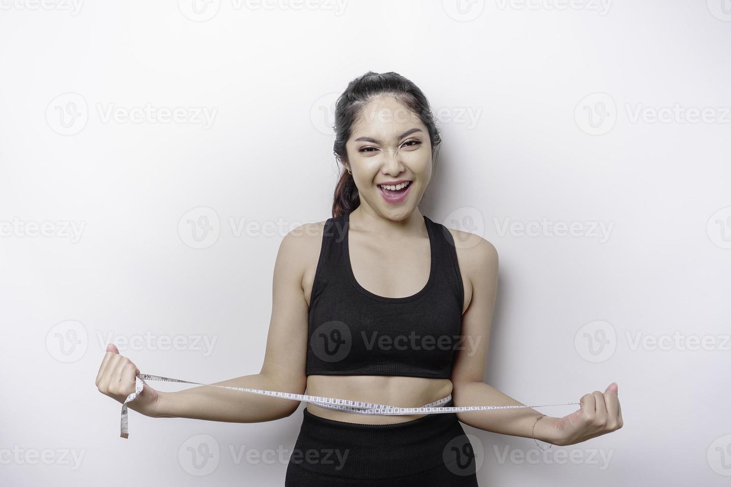 porträt einer fröhlichen jungen asiatischen frau in sportbekleidung, die die taille mit band misst und sich glücklich fühlt, zu hause gewicht zu verlieren. gesunder lebensstil, sport und schlankheitsdiätkonzept foto