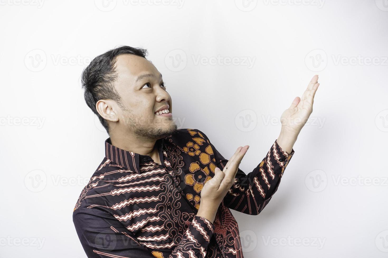 Aufgeregter asiatischer Mann mit Batikhemd, der auf den Kopierraum auf ihm zeigt, isoliert durch weißen Hintergrund foto