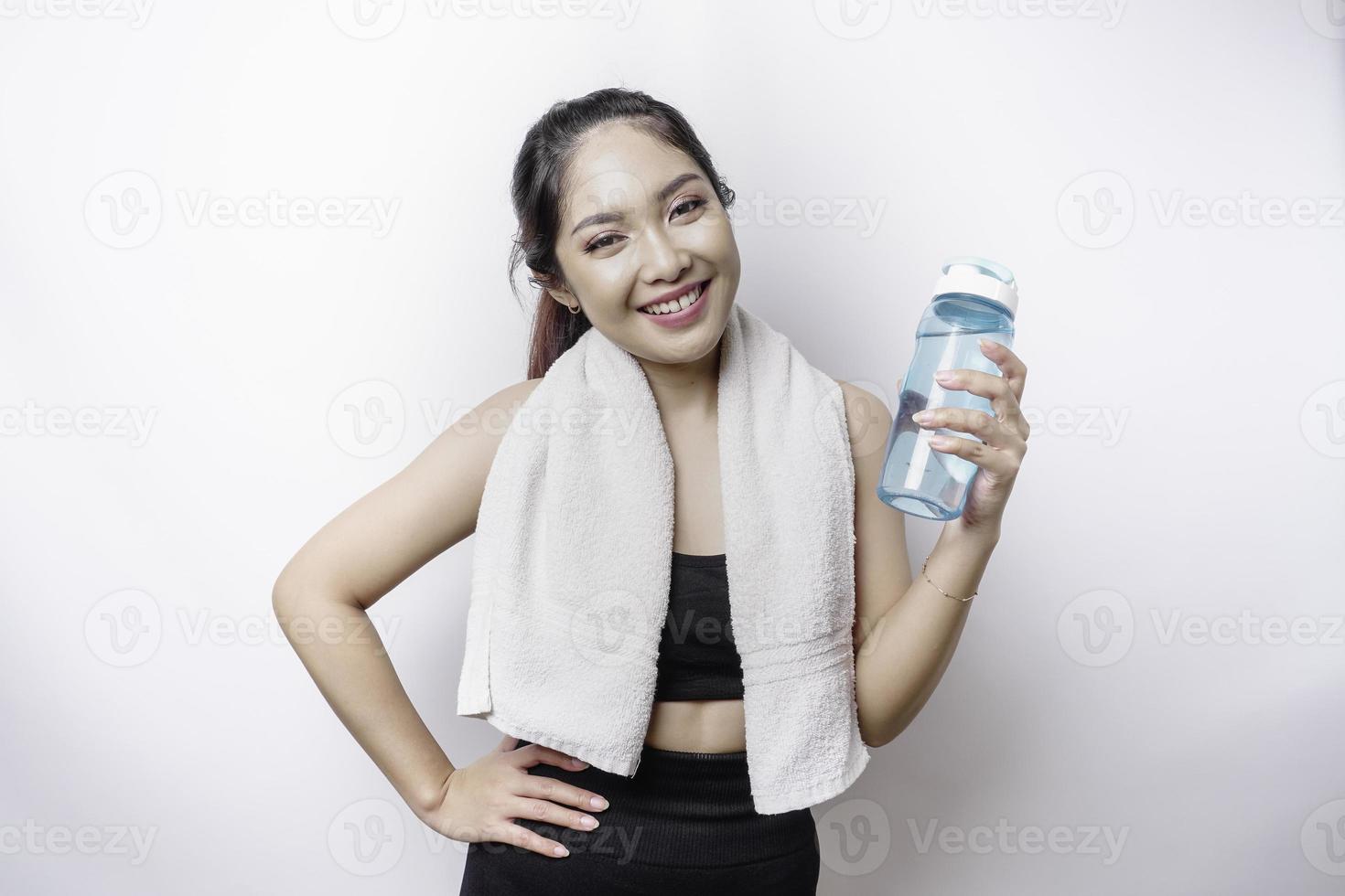 sportliche asiatin posiert mit einem handtuch auf der schulter und hält eine flasche wasser, lächelt und entspannt sich nach dem training foto