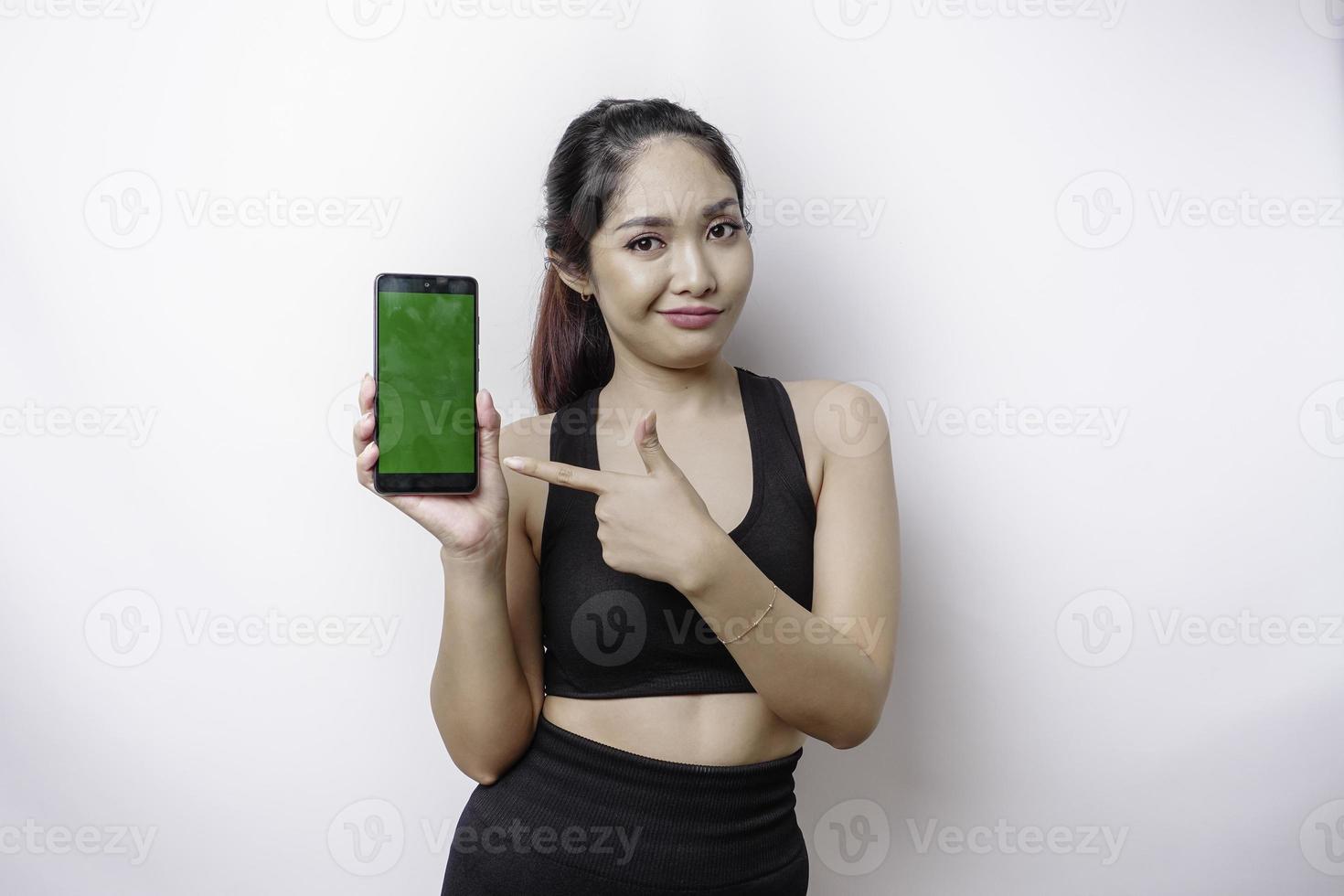 eine nachdenkliche junge sportliche frau trägt sportbekleidung, während sie ihr kinn hält und einen grünen bildschirm auf ihrem telefon zeigt, isoliert durch weißen hintergrund foto