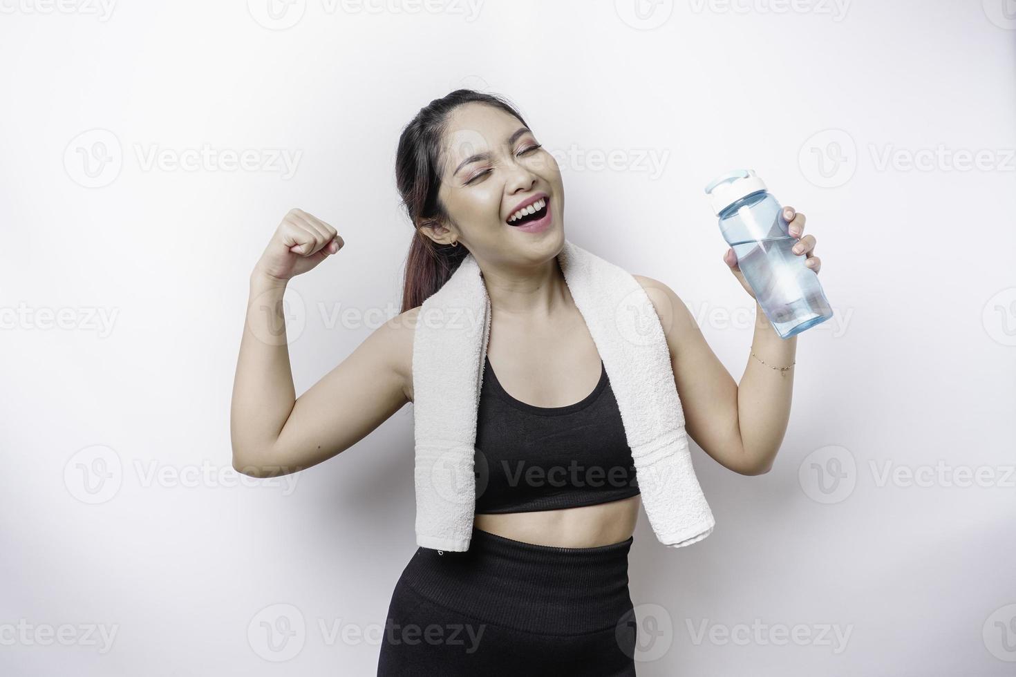 sportliche asiatische frau, die mit einem handtuch auf ihrer schulter posiert und eine flasche wasser hält, die eine starke pose gestikuliert, indem sie ihren arm hebt foto