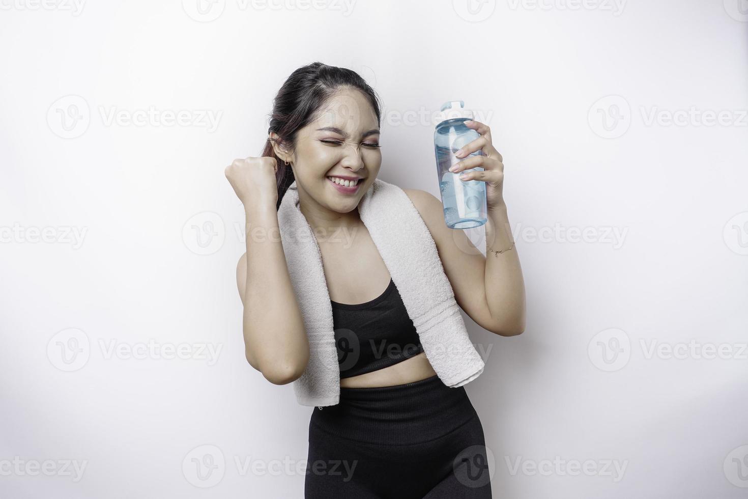 lächelnde erfolgreiche sportliche asiatische frau, die mit einem handtuch auf ihrer schulter posiert und eine flasche wasser hält foto