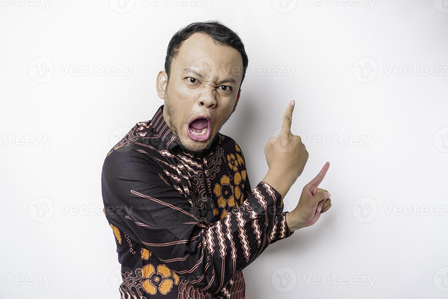 ernsthafter asiatischer Mann mit Batikhemd, der auf den Kopierraum auf ihm zeigt, isoliert durch weißen Hintergrund foto
