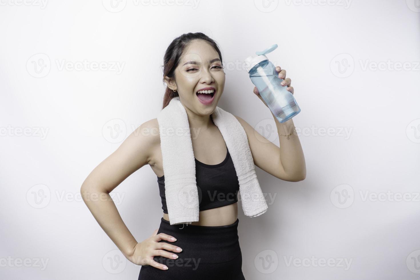 sportliche asiatin posiert mit einem handtuch auf der schulter und hält eine flasche wasser, lächelt und entspannt sich nach dem training foto