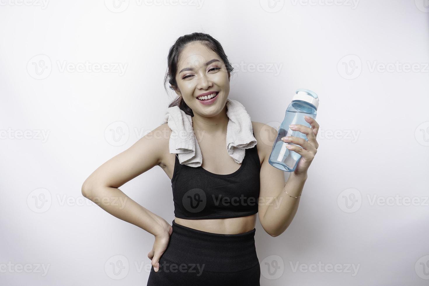 sportliche asiatin posiert mit einem handtuch auf der schulter und hält eine flasche wasser, lächelt und entspannt sich nach dem training foto