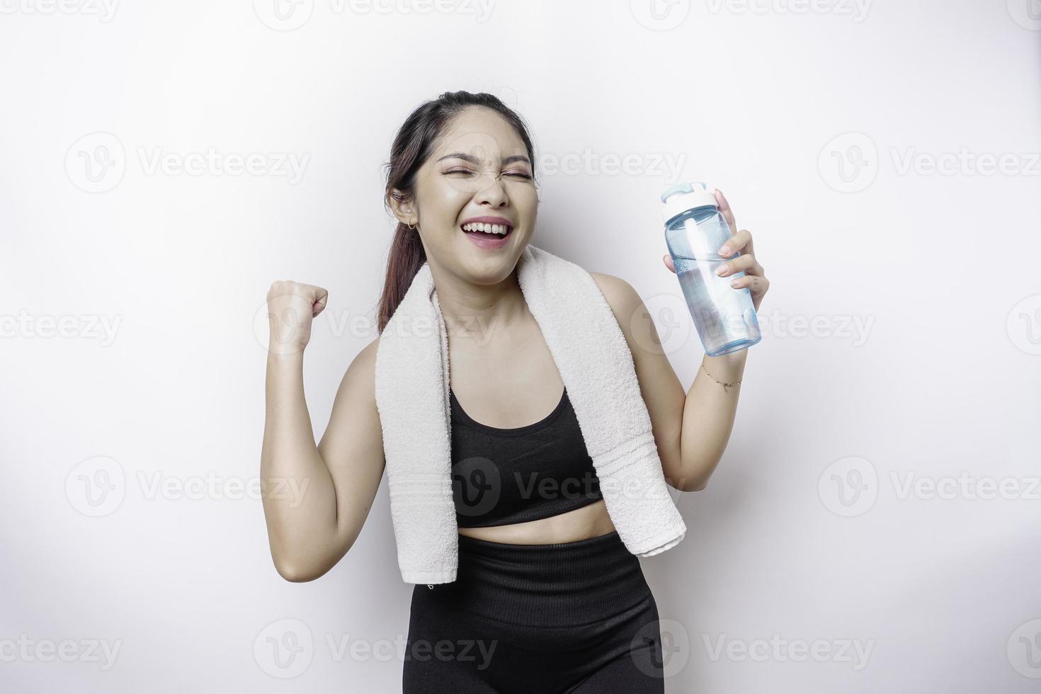 lächelnde erfolgreiche sportliche asiatische frau, die mit einem handtuch auf ihrer schulter posiert und eine flasche wasser hält foto