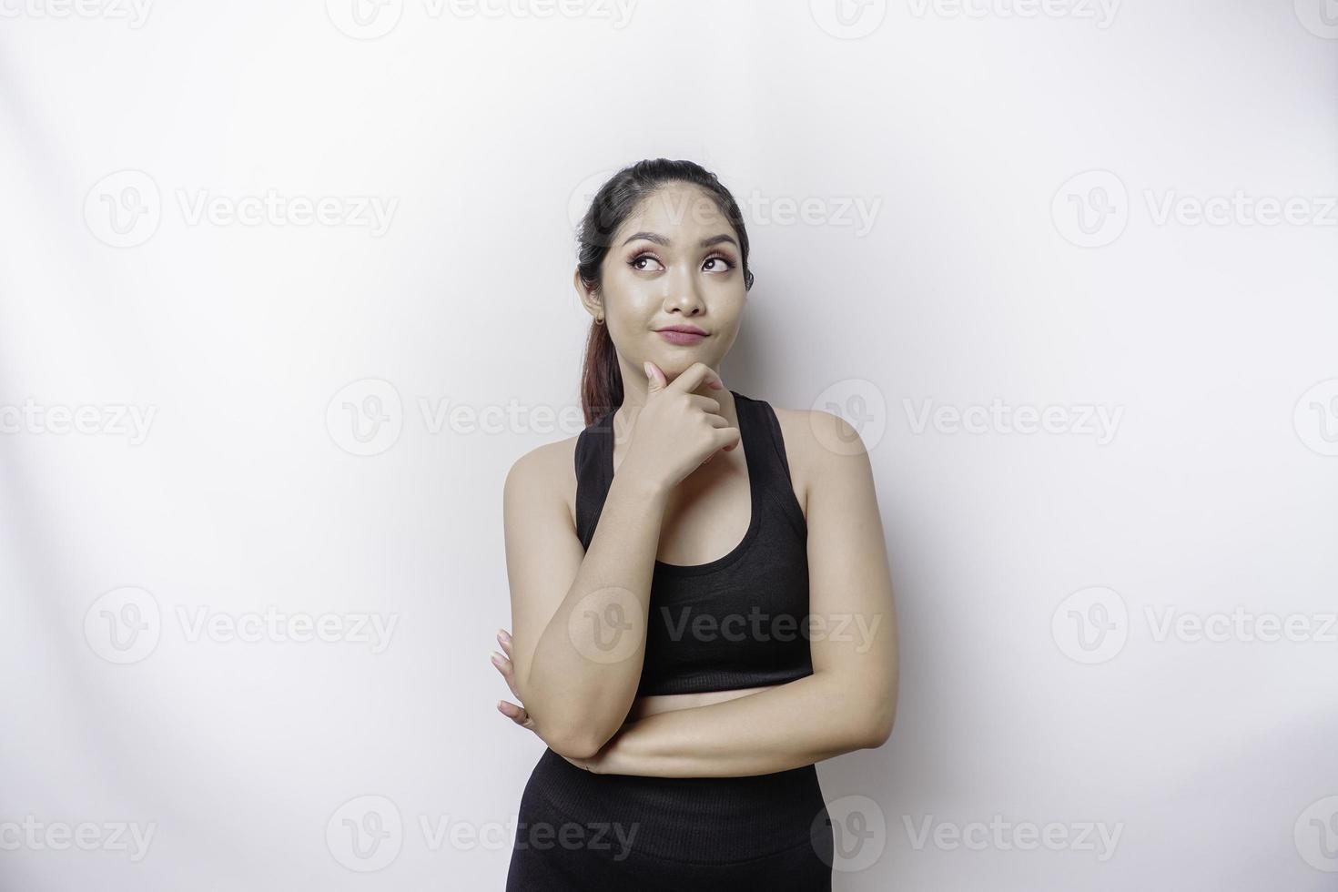 nachdenkliche sportliche asiatische frau, die sportkleidung trägt, schaut beiseite, während sie ihr kinn berührt, isoliert durch weißen hintergrund. foto