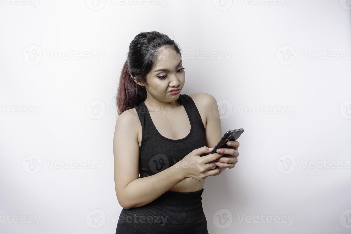 eine unzufriedene sportliche asiatin sieht verärgert aus und trägt gereizte gesichtsausdrücke von sportbekleidung, die ihr telefon halten foto