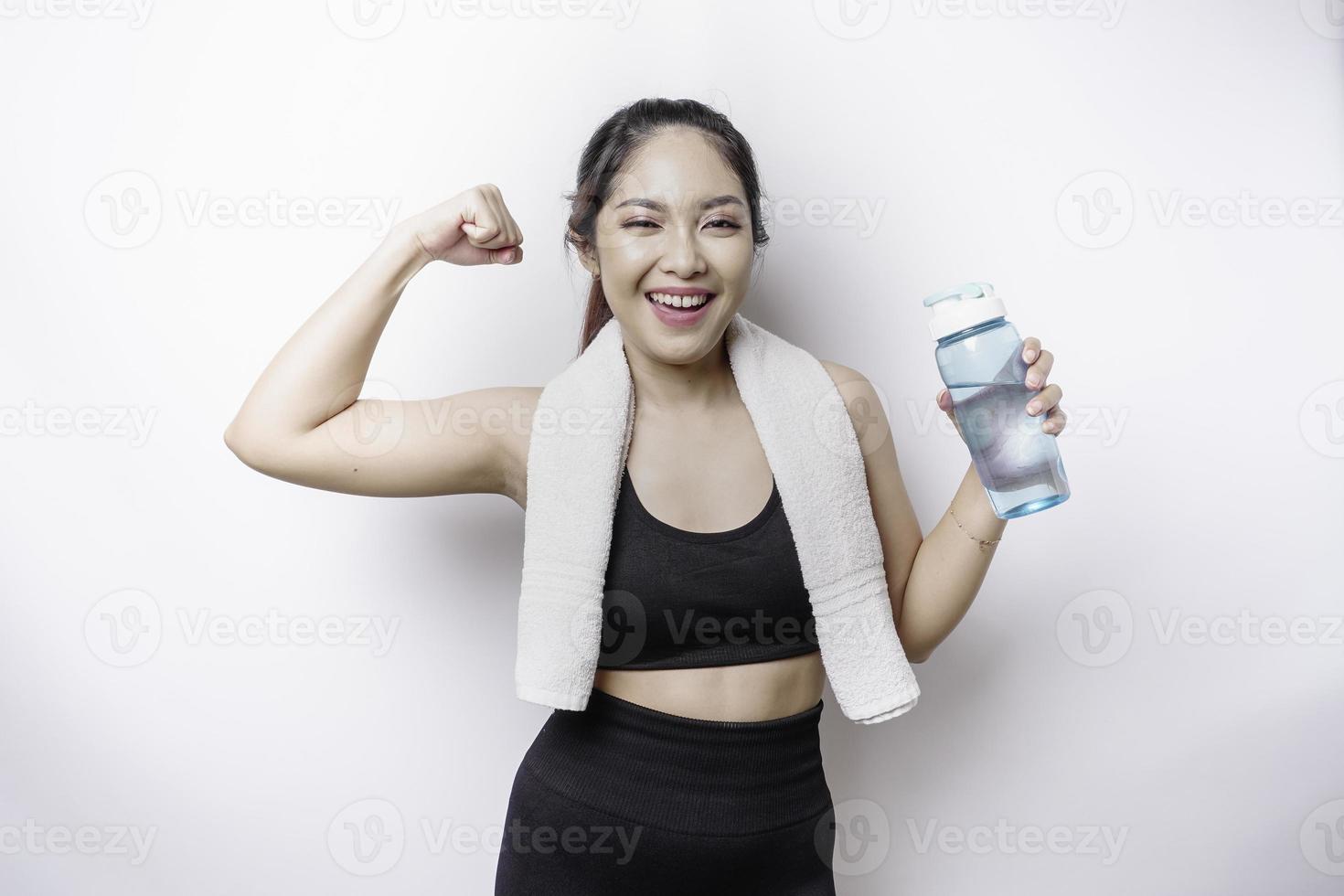 sportliche asiatische frau, die mit einem handtuch auf ihrer schulter posiert und eine flasche wasser hält, die eine starke pose gestikuliert, indem sie ihren arm hebt foto