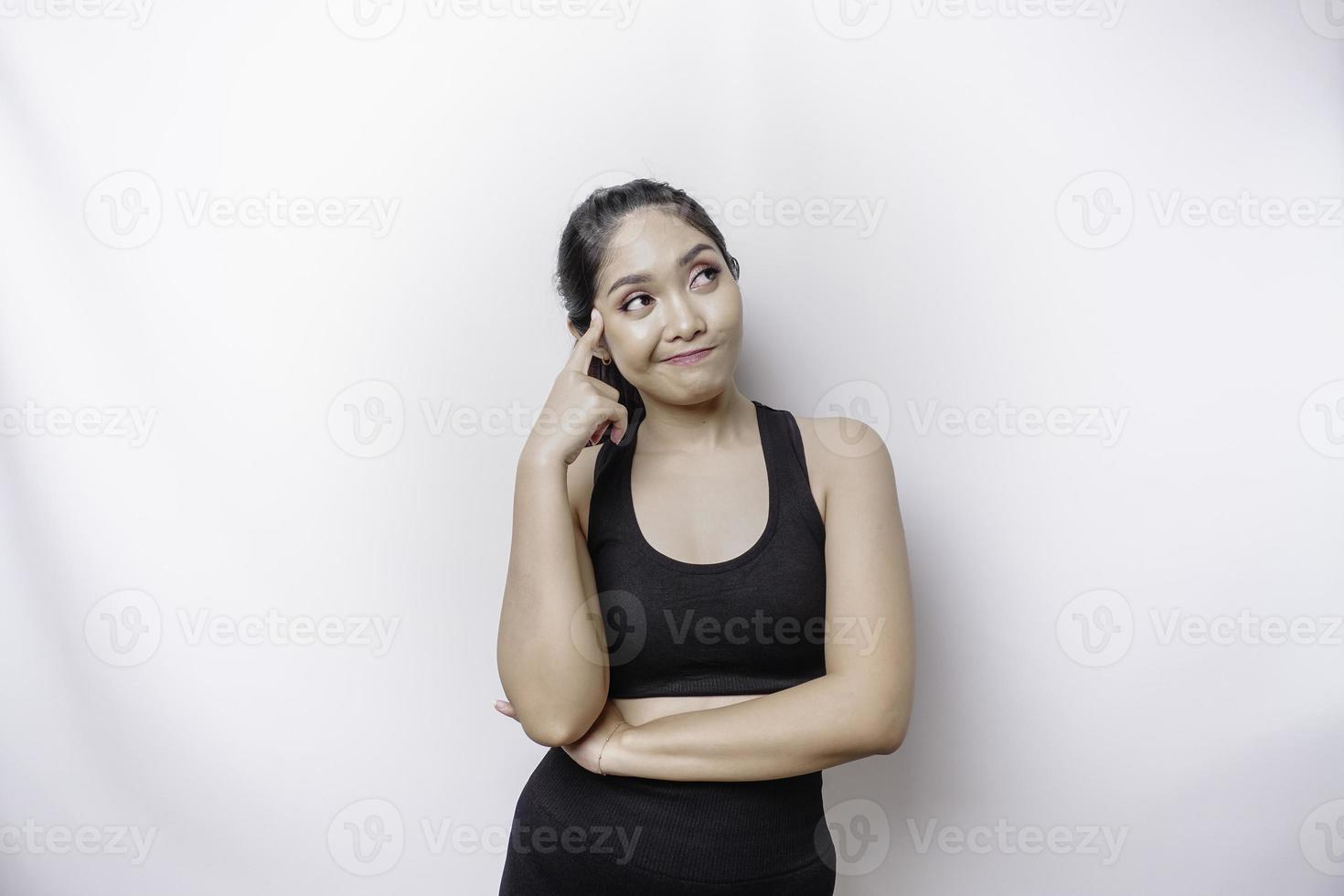 nachdenkliche sportliche asiatische frau, die sportkleidung trägt, schaut beiseite, während sie ihr kinn berührt, isoliert durch weißen hintergrund. foto