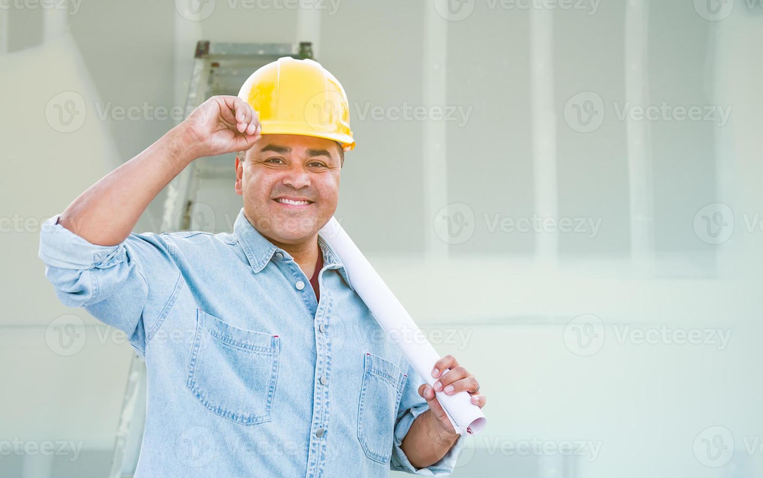 Hispanischer männlicher Auftragnehmer mit Blaupausenplänen mit Schutzhelm vor Trockenbau und Leiter foto