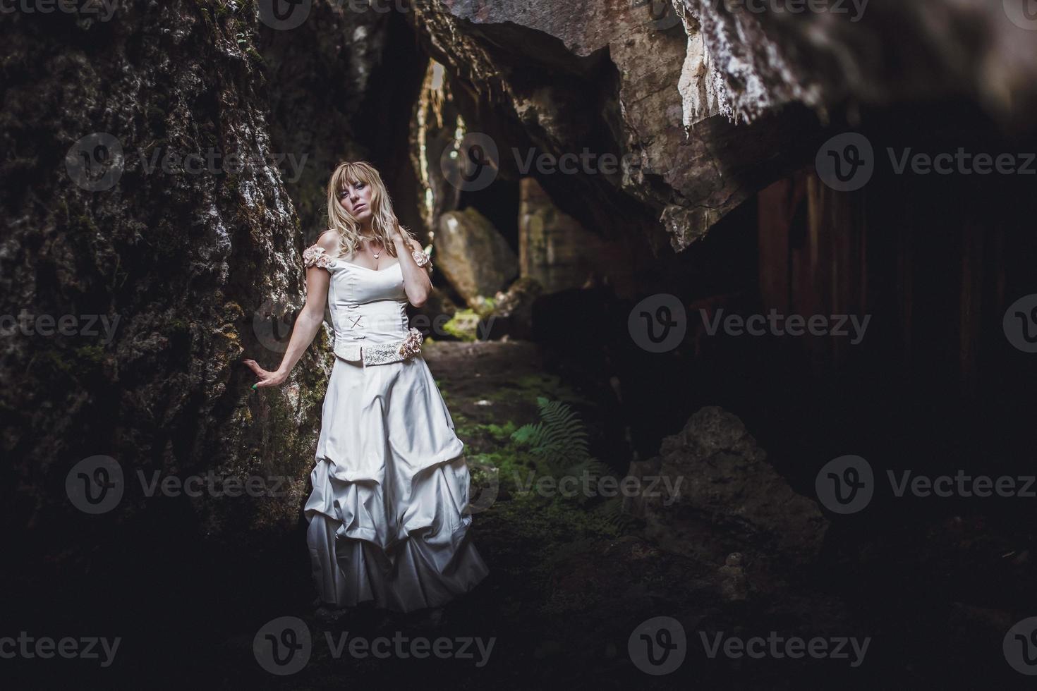 Schönes blondes Mädchen in einem weißen Kleid mit einem Korsett in einer tiefen dunklen Höhle foto