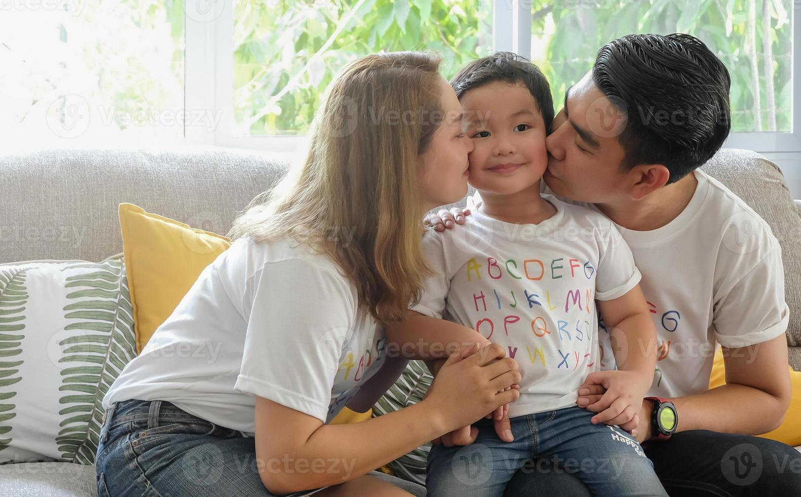 junge asiatische eltern küssen ihren kleinen sohn mit liebe am sofa wohnzimmer wärmehaus hintergrund, lichteffekt und kopierraum. foto