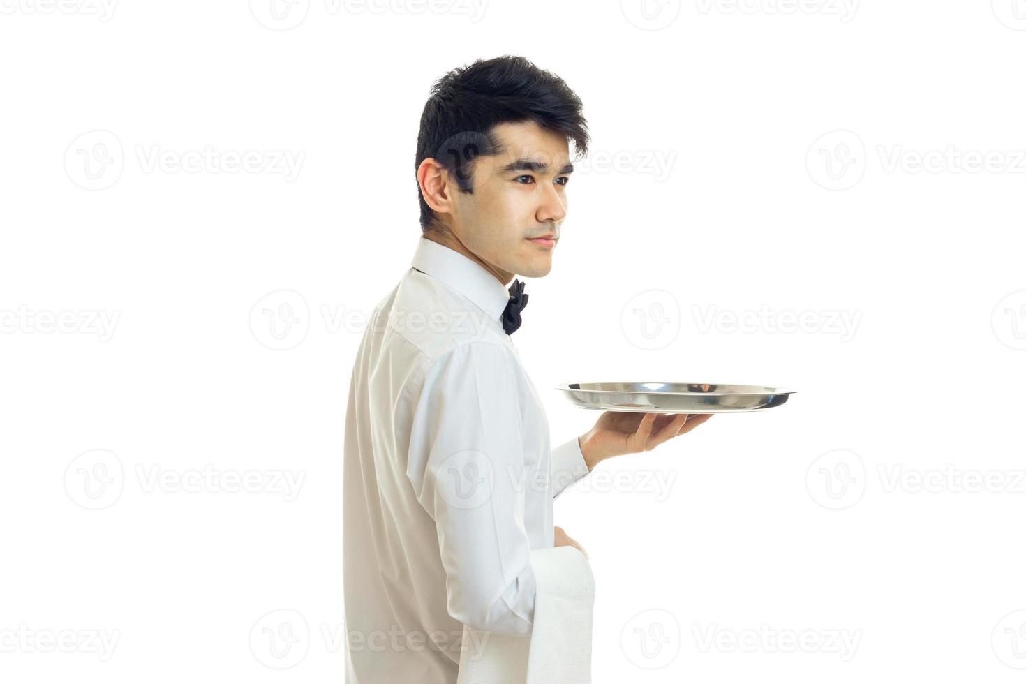 der junge kellner mit schwarzen haaren und weißem hemd steht seitwärts und hält das leere tablett in der hand foto
