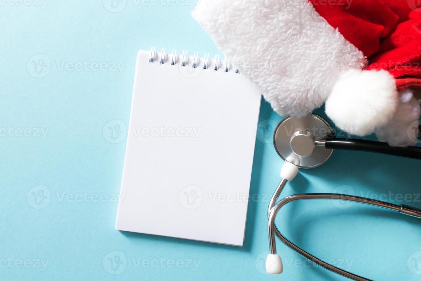 medizinische weihnachts- und neujahrskomposition mit stethoskop, notizblock und weihnachtsmütze auf blauem hintergrund. gesundheits- und medizinkonzept für karte, kalender, cover. foto