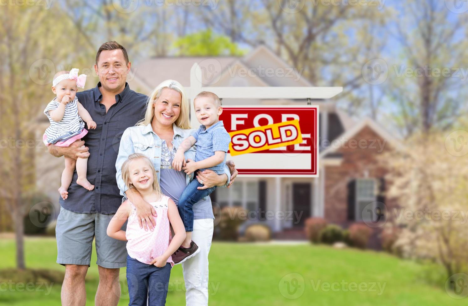 glückliche junge kaukasische familie draußen vor ihrem neuen zuhause und verkauftem immobilienschild foto