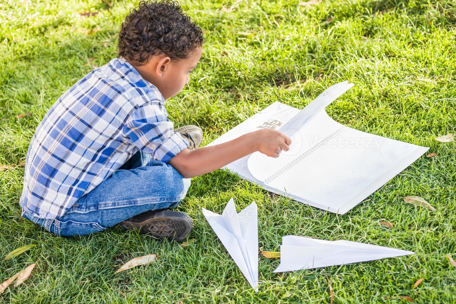 afroamerikanischer und mexikanischer junge, der lernt, wie man papierflugzeuge draußen auf dem gras faltet foto