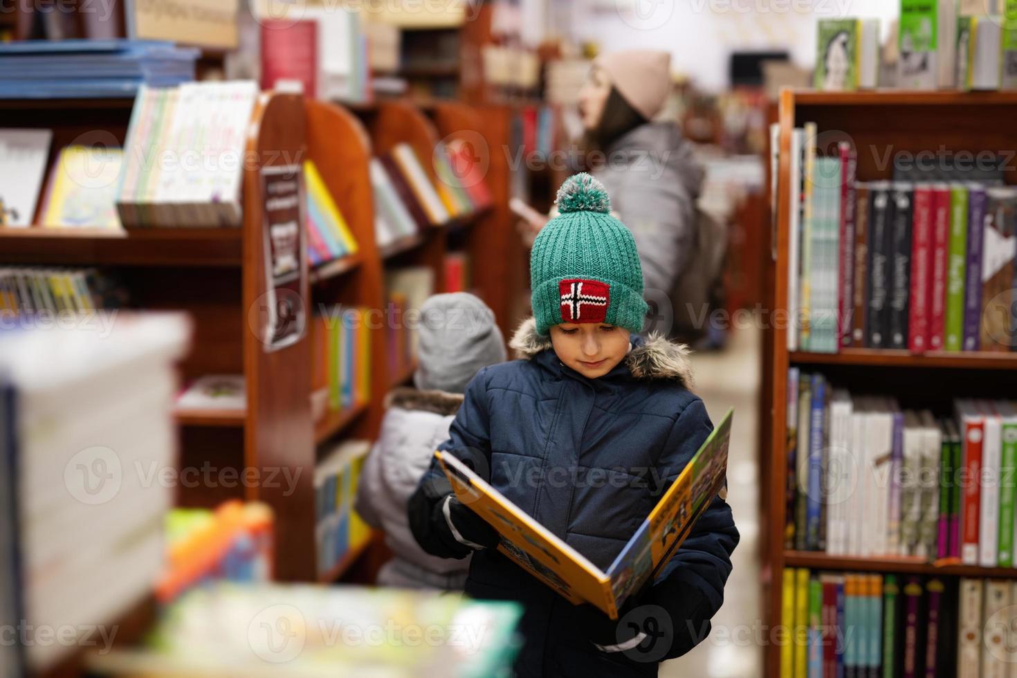 Junge in Jacke und Hut, der ein Buch aus dem Bücherregal in der Bibliothek erreicht. Lernen und Bildung europäischer Kinder. foto