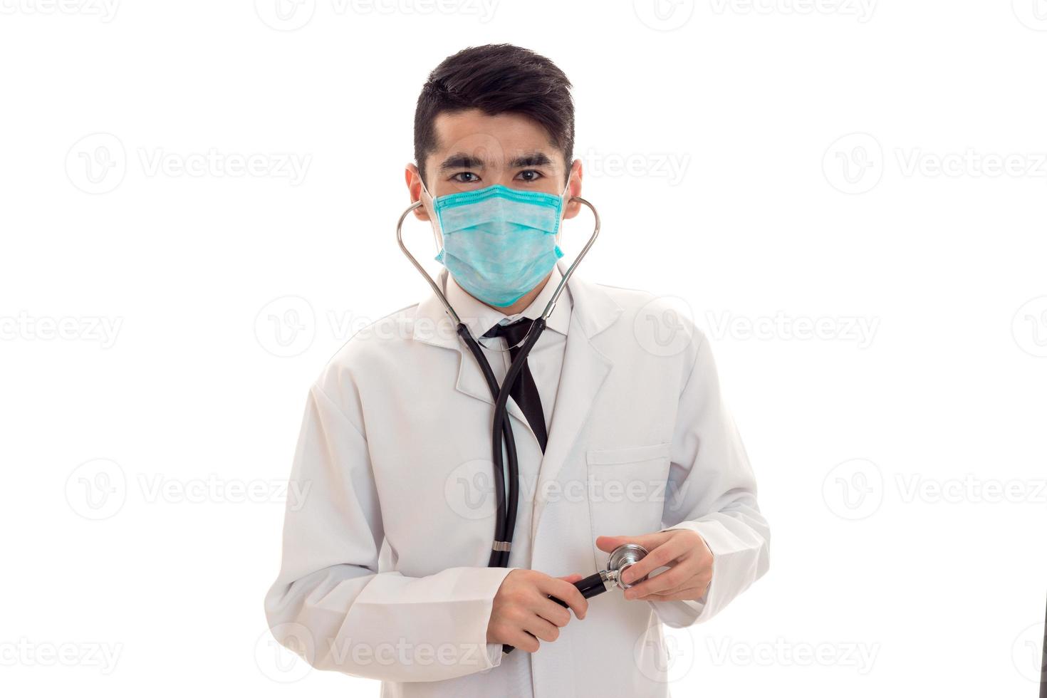 junger männlicher Arzt in Uniform und Maske mit Stethoskop, der isoliert auf weißem Hintergrund in die Kamera schaut foto