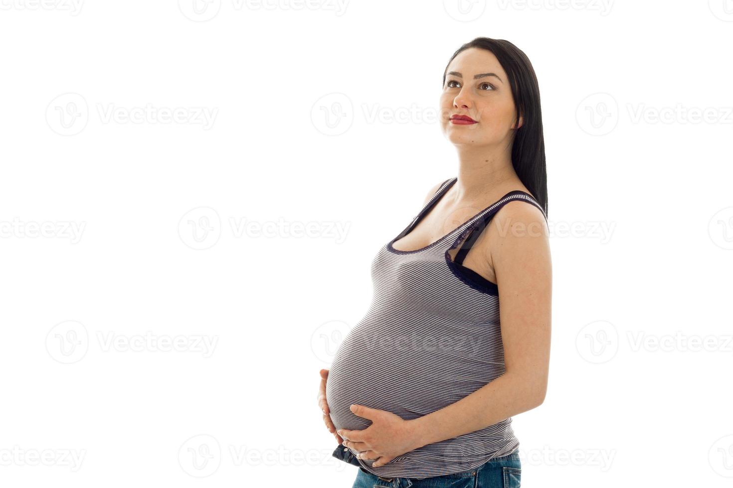 schöne brünette schwangere Frau, die ihren Bauch berührt und isoliert auf weißem Hintergrund nach oben schaut foto