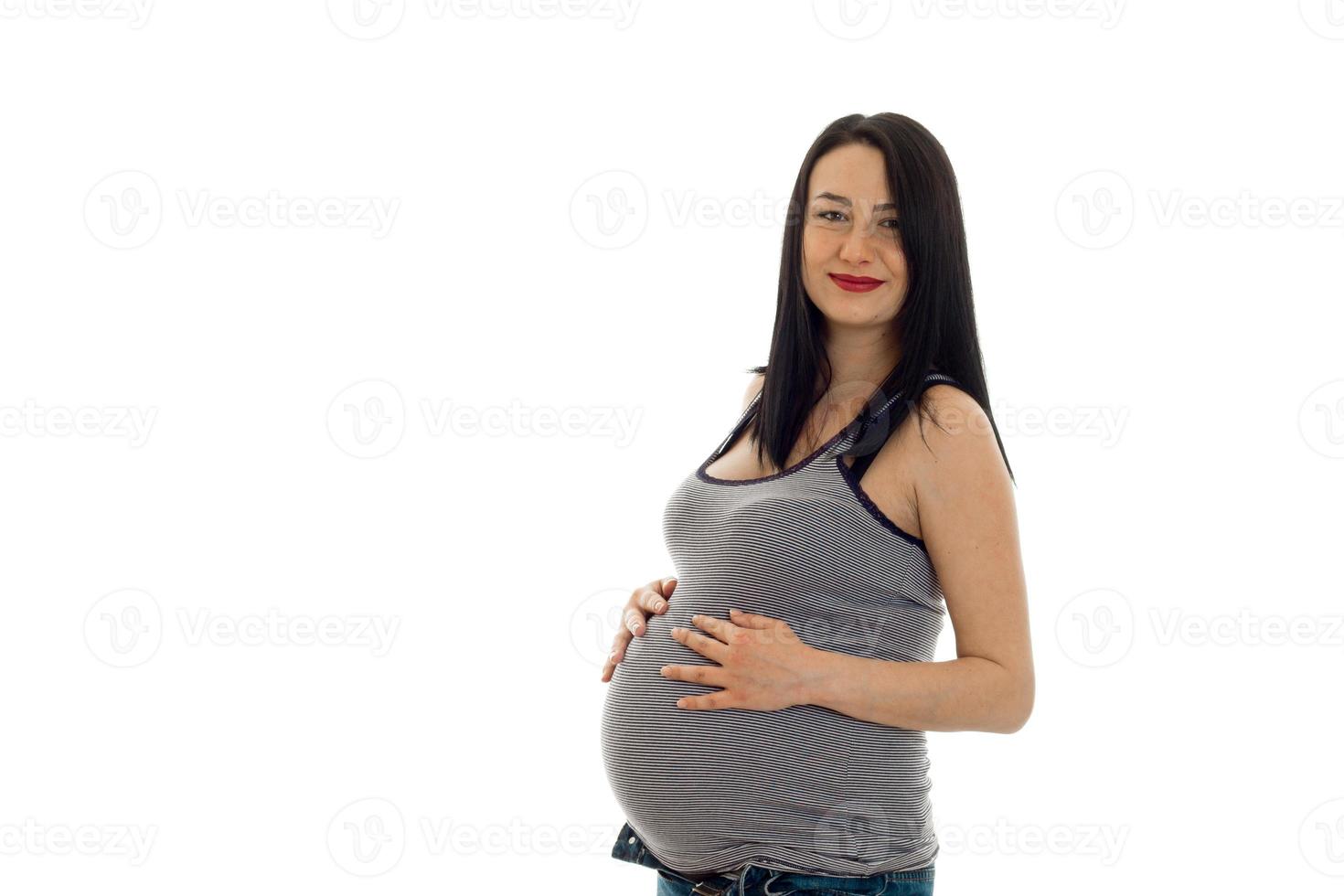 Junges schwangeres Mädchen mit dunklem Haar, das ihren dicken Bauch berührt und isoliert auf weißem Hintergrund in die Kamera schaut foto