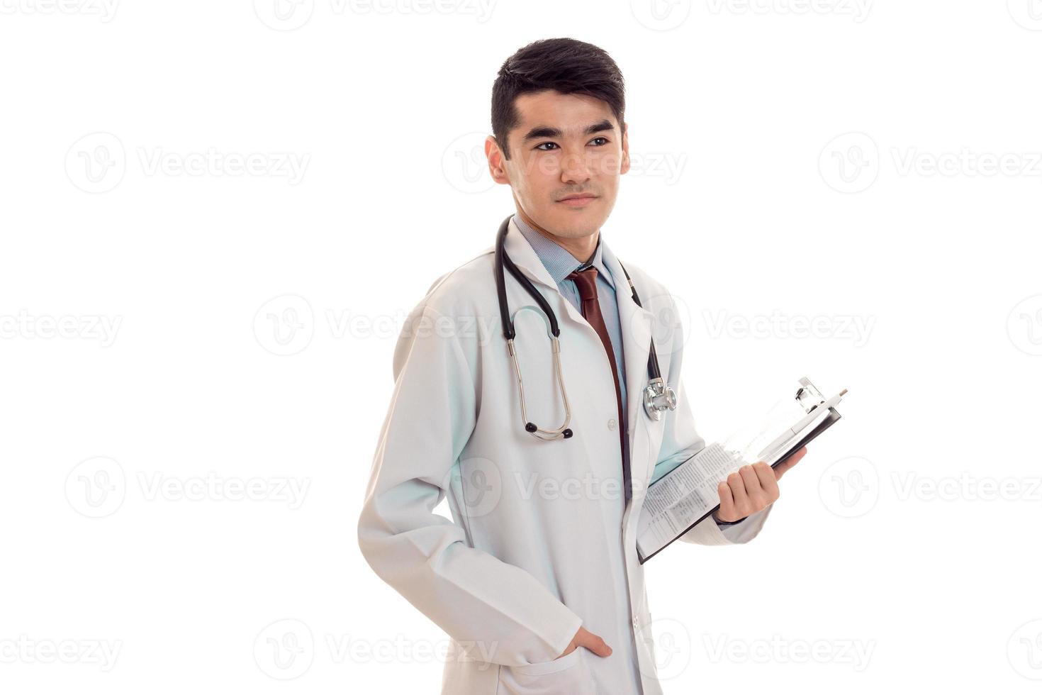 nachdenklicher hübscher brünetter männlicher Arzt in Uniform mit Stethoskop am Hals posiert im Studio isoliert auf weißem Hintergrund foto