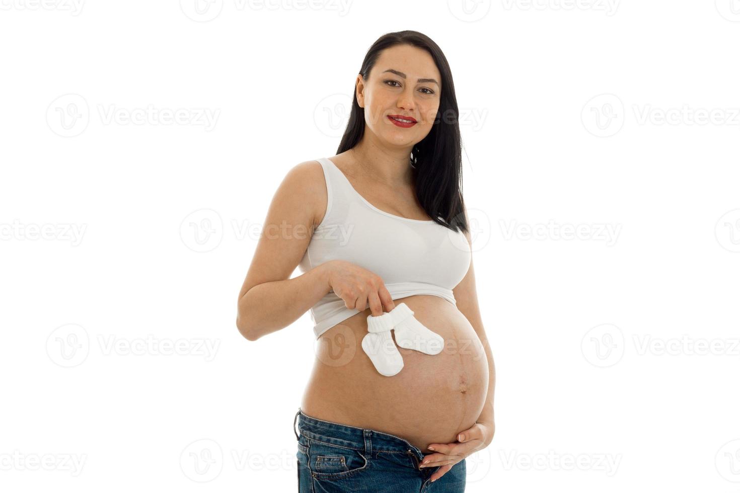 Junges schwangeres Mädchen mit dunklen Haaren, das ihren dicken Bauch mit kleinen weißen Socken in den Händen berührt und isoliert auf weißem Hintergrund in die Kamera schaut foto