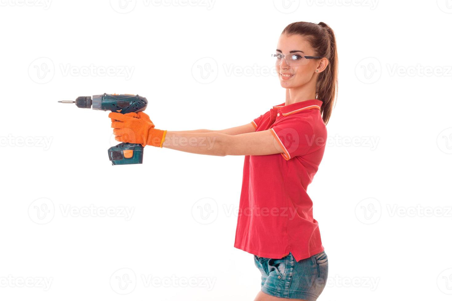 Junge fröhliche brünette Baumeisterin in Uniform mit Brille und Bohrer in ihren Händen machen Reovations isoliert auf weißem Hintergrund foto