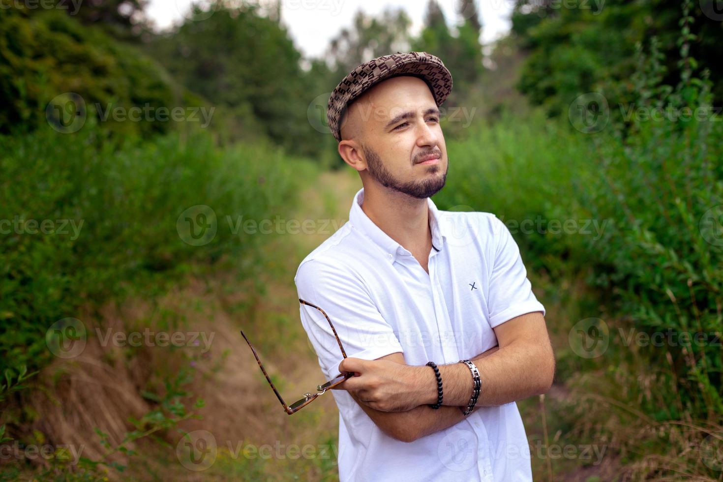 Wunderschöner Mann mit Hut hält eine Sonnenbrille in der Hand und blickt auf den grünen Park foto