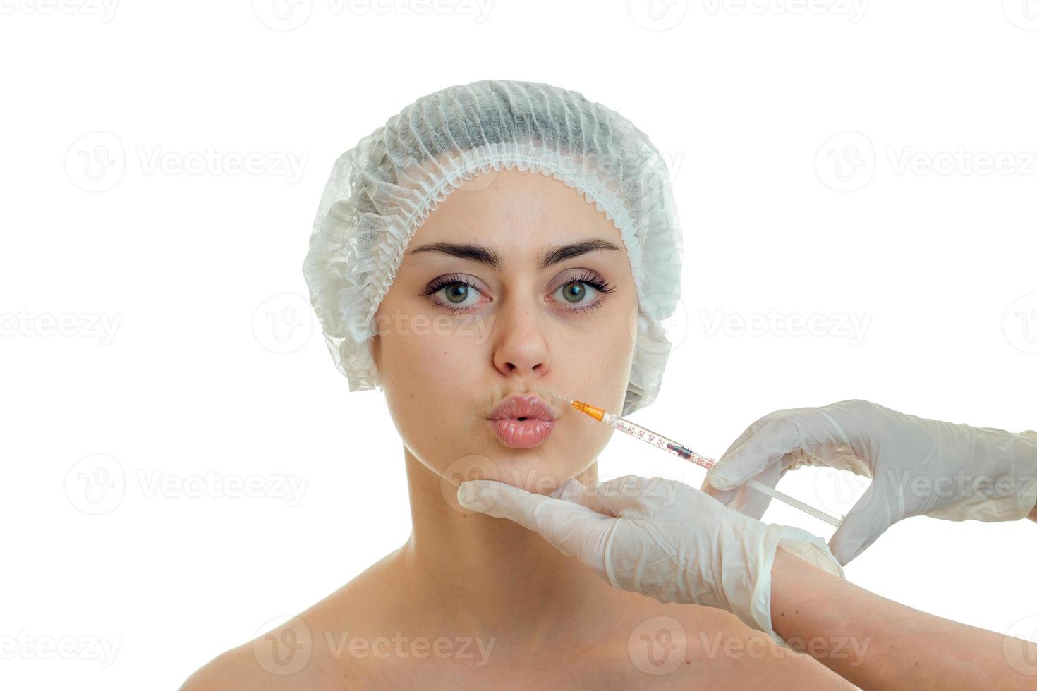junges schönes mädchen in medizinischem haarhut sieht gerade aus, macht lippenküsse und der arzt setzt ihr eine spritze ins gesicht foto