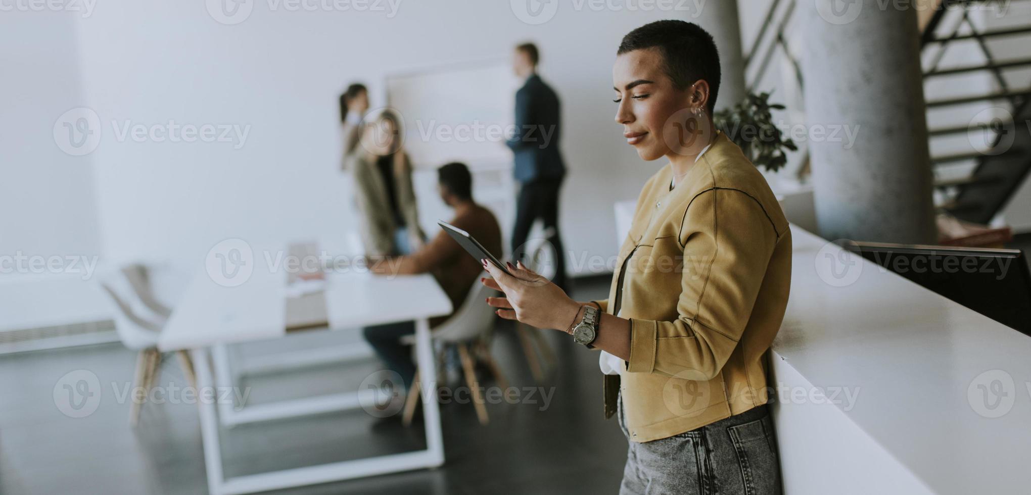 Junge Geschäftsfrau mit kurzen Haaren, die im Büro steht und vor ihrem Team ein digitales Tablet verwendet foto