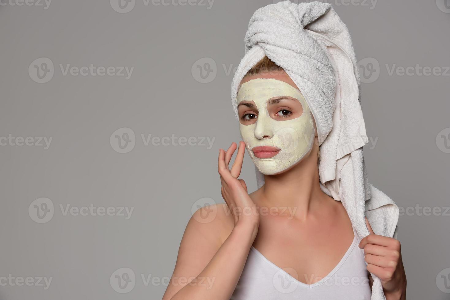 Schönes weibliches Modell mit Handtuch auf ihrem Ohr und weißer kosmetischer Gesichtsmaske auf ihrem Gesicht. Beauty-Kosmetik-Konzept. isoliert auf grauem Hintergrund. foto