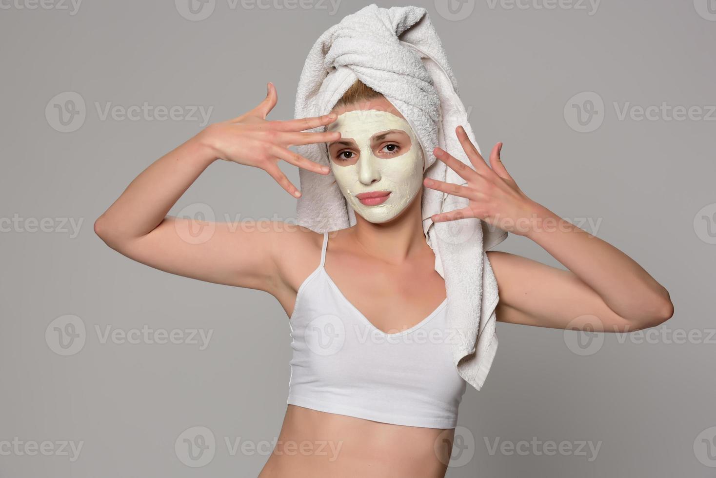 Schönes weibliches Modell mit Handtuch auf ihrem Ohr und weißer kosmetischer Gesichtsmaske auf ihrem Gesicht. Beauty-Kosmetik-Konzept. isoliert auf grauem Hintergrund. foto