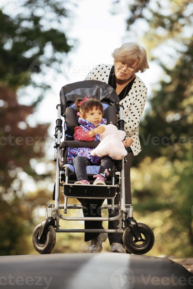 glückliche ältere Dame, die Rollstuhl und Kinder schiebt. Großmutter und Kinder genießen einen Spaziergang im Park. Kind, das behinderte Großeltern unterstützt. Familienbesuch. Generationen Liebe und Beziehung foto
