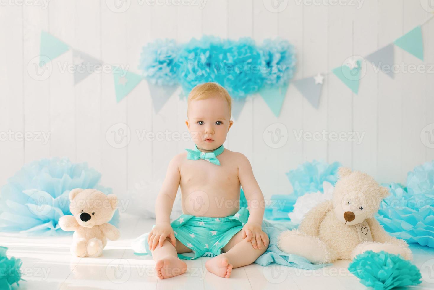 ein Baby sitzt neben kuscheligen Teddybären in festlicher Geburtstagsdeko, ein einjähriger Junge in Blau mit blauen Girlanden foto